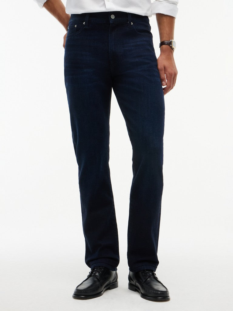 A person wearing dark navy blue jeans, a white shirt, and black leather dress shoes, shown from the waist down against a plain white studio background.