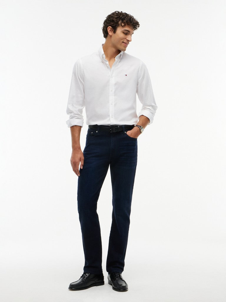 A man modeling a white long-sleeve button-down shirt and dark navy blue jeans against a plain white studio background.