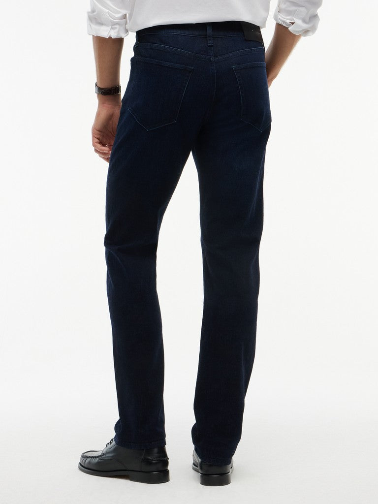Rear view of a person wearing dark navy blue jeans, a white shirt, and black shoes against a plain white studio background.