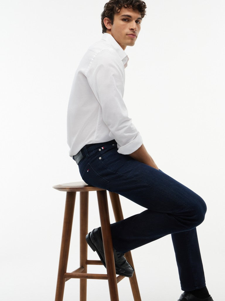 A man wearing a white long-sleeve button-down shirt and dark navy blue jeans sits on a wooden stool against a plain white studio background.