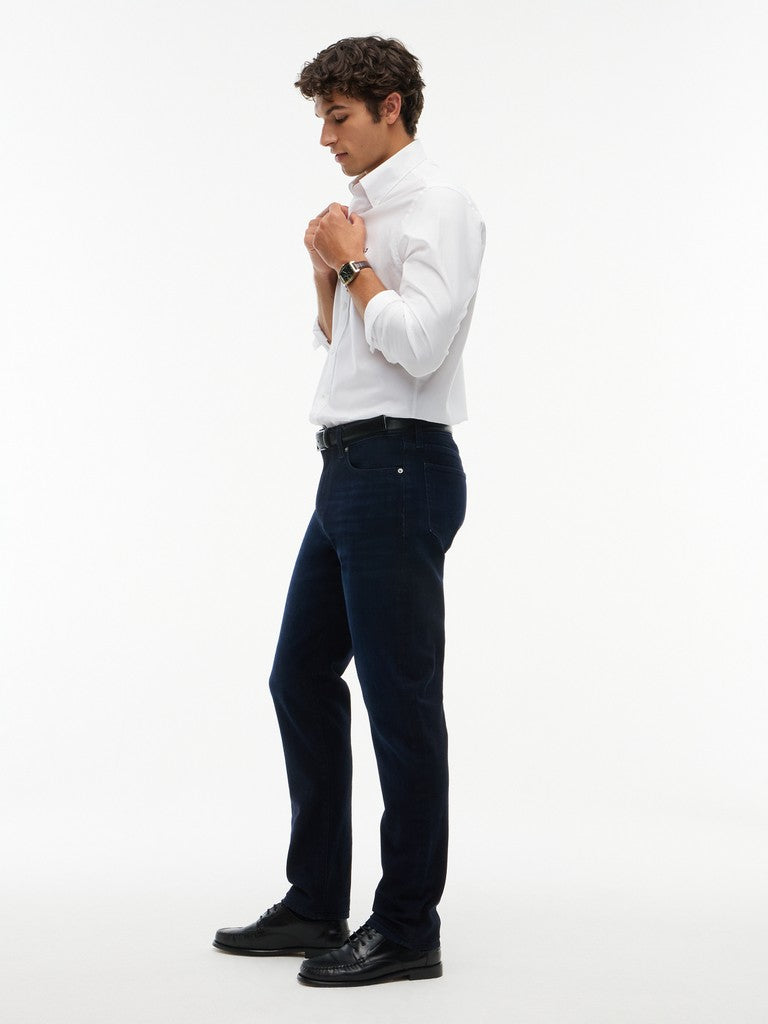 A man wearing a white long-sleeve dress shirt, dark navy jeans, and black dress shoes, standing in profile against a plain white studio background.