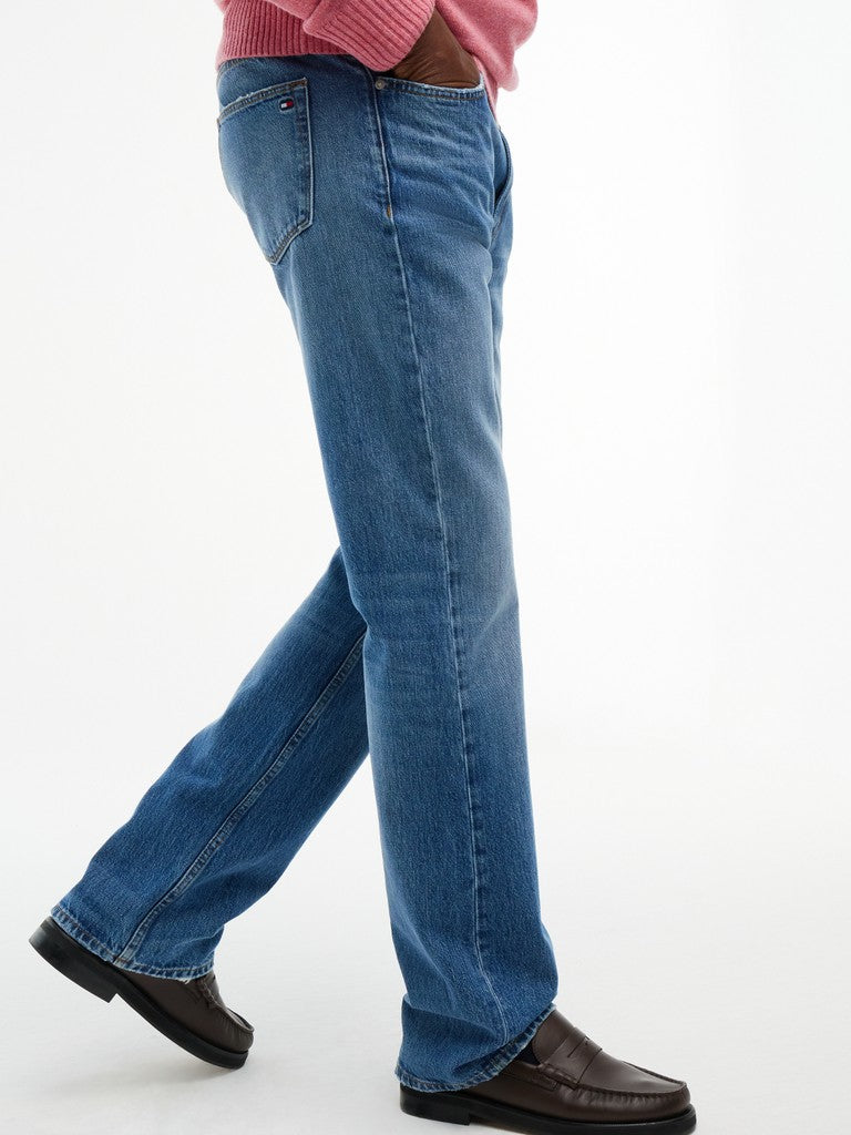 A person wearing medium blue denim jeans with a pink ribbed top and dark brown leather loafers, shown from the waist down in a side-profile walking pose against a plain white studio background.