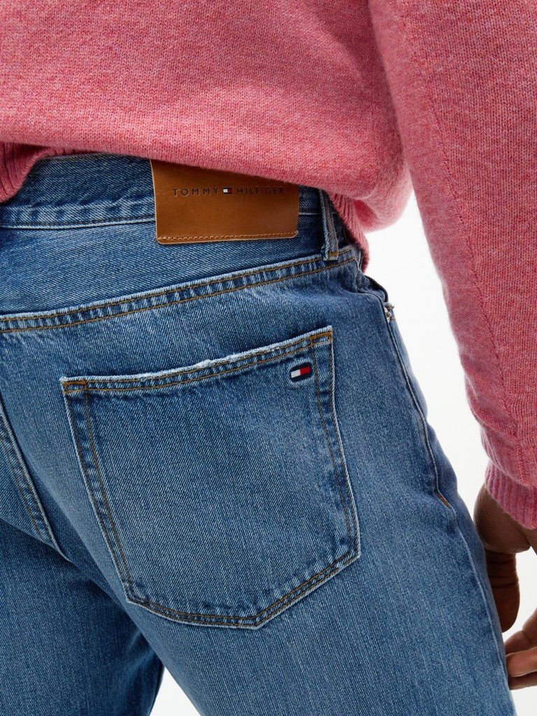 Close-up of the back waistband and pocket area of blue denim jeans worn by a person, with a pink heathered sweater visible at the top and a small Tommy Hilfiger flag logo on the pocket.