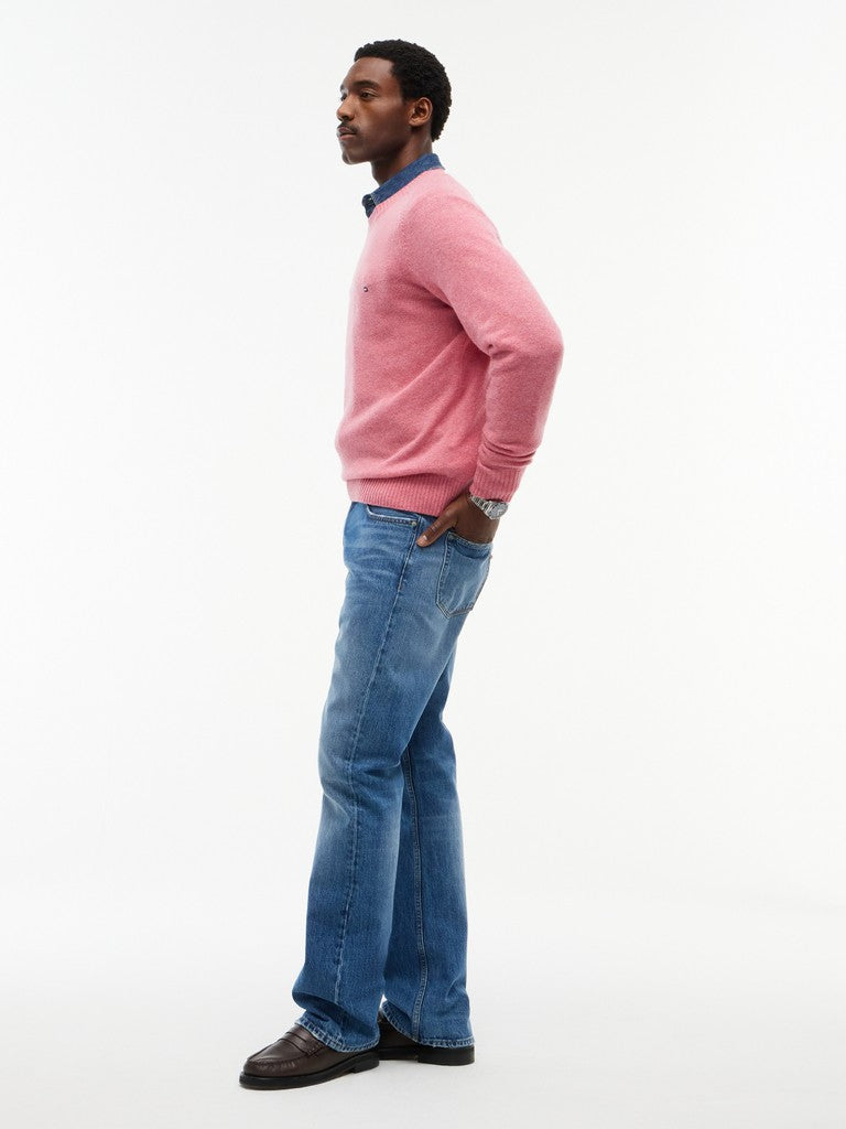 A man in profile wearing a pink knit sweater over a blue collared shirt, blue jeans, and dark brown shoes, standing against a plain white studio background.