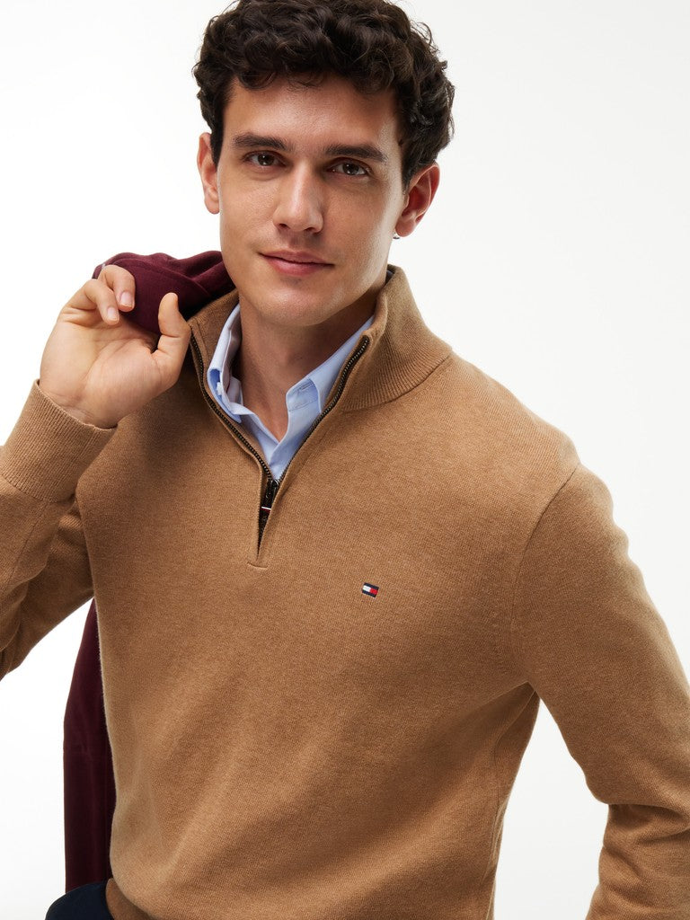 A man wearing a camel brown quarter-zip sweater over a light blue dress shirt, holding a dark burgundy scarf against a plain white background. The Tommy Hilfiger flag logo is visible on the chest.