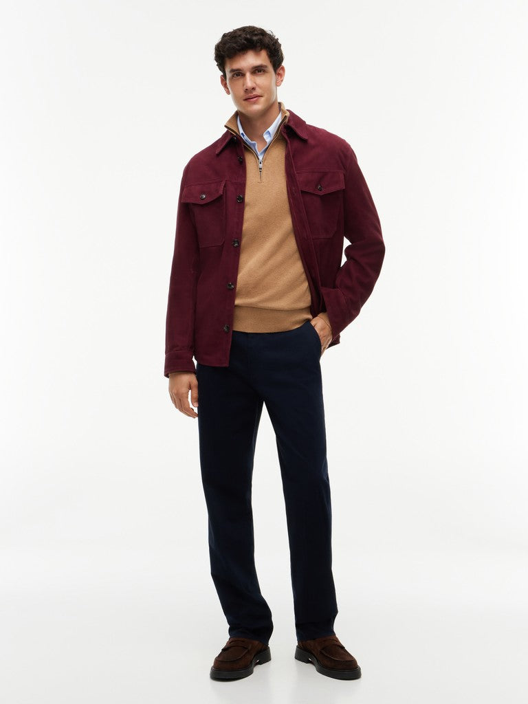 A man modeling a burgundy overshirt jacket over a tan quarter-zip sweater, a light blue patterned collared shirt, dark navy trousers, and brown suede shoes, photographed against a seamless white studio background.