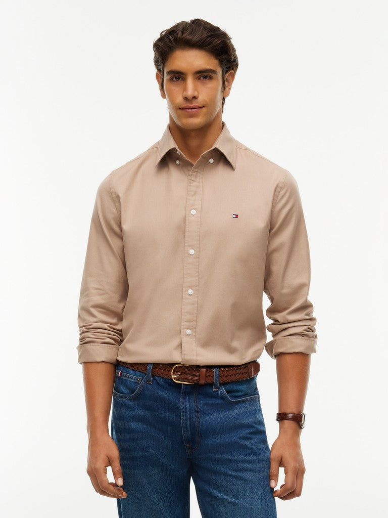 A man wearing a light beige long-sleeve button-down shirt with rolled-up sleeves, paired with blue jeans and a brown belt, photographed against a plain white studio background.
