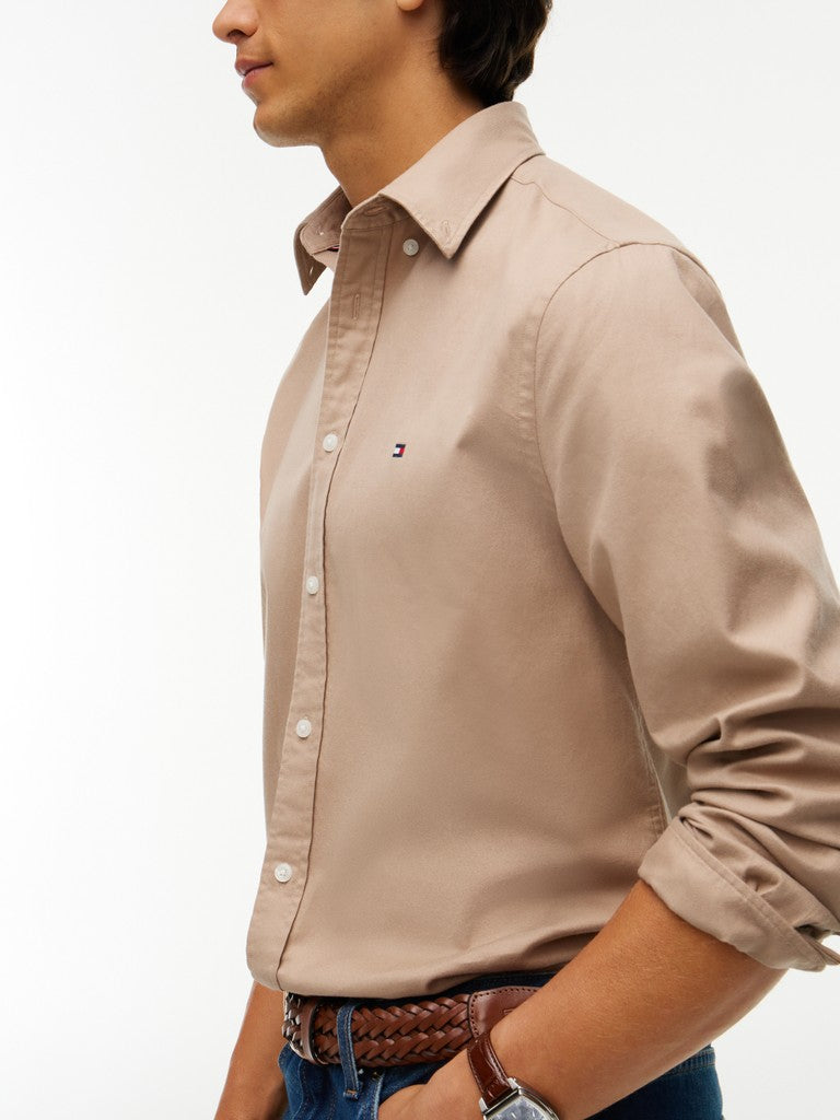 A man wearing a light beige long-sleeve button-down shirt with rolled-up sleeves, paired with a brown braided belt and blue jeans, shown in a side profile against a white background.