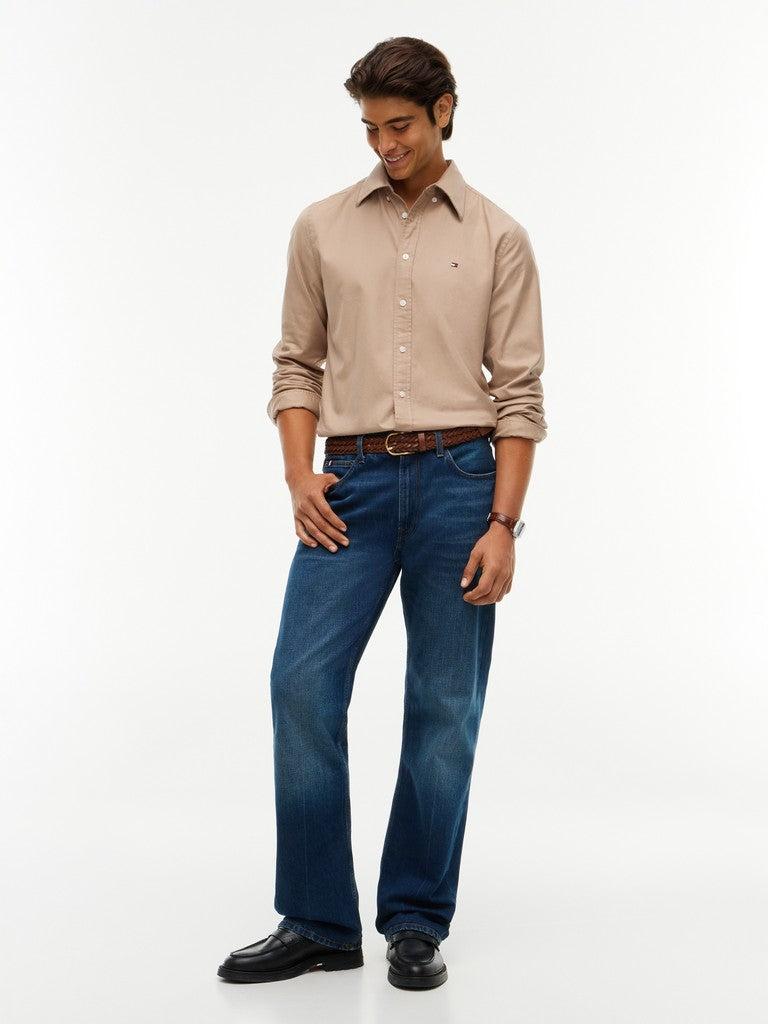 A man modeling a beige long-sleeve button-down shirt and blue jeans with a brown belt, standing against a plain white studio background.