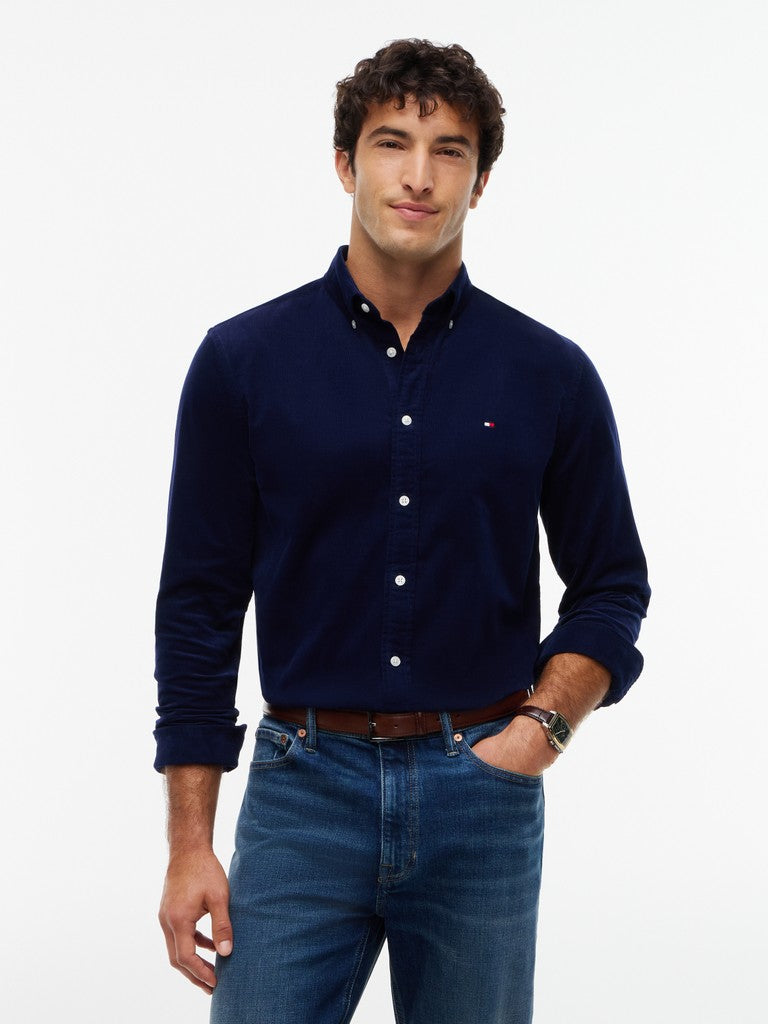 A man wearing a Tommy Hilfiger navy blue long-sleeve button-down shirt and blue jeans, standing against a plain white studio background.