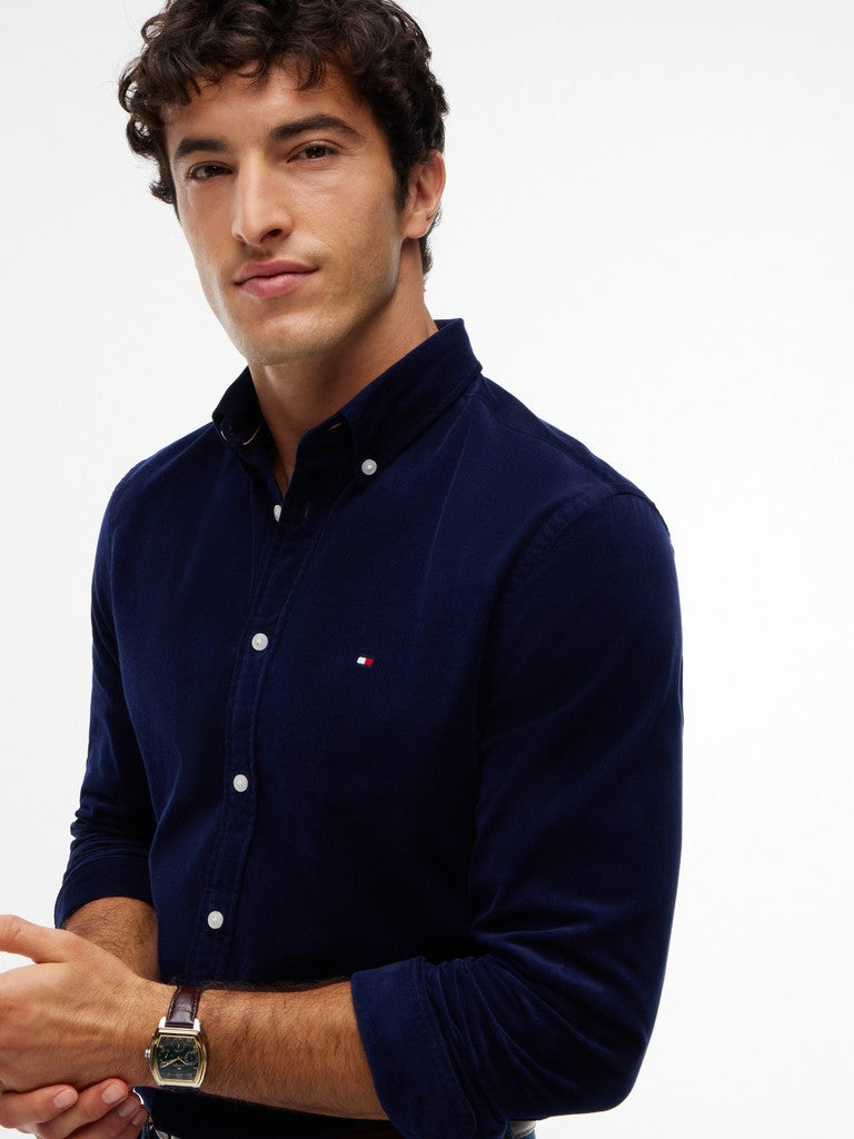 A man wearing a dark navy blue long-sleeve button-down shirt with white buttons and a small red and white embroidered logo on the chest, photographed against a plain white background.