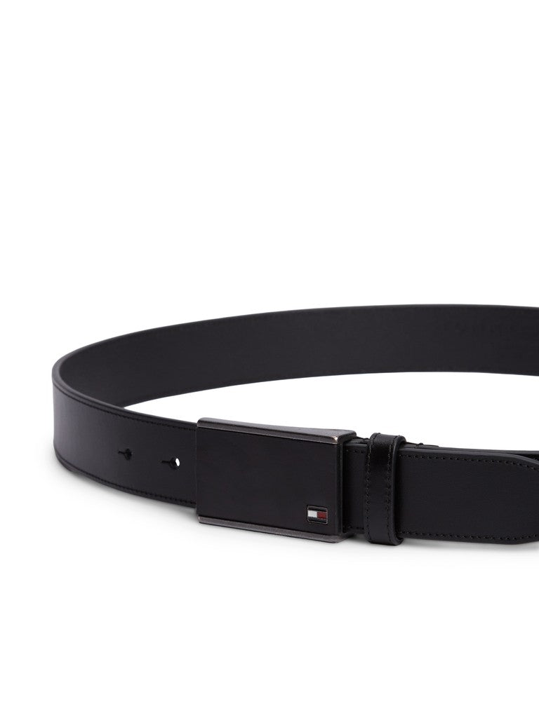 Black leather belt on a white studio surface The belt is fastened with a black metallic buckle with a subtle Hilfiger flag logo in the bottom corner