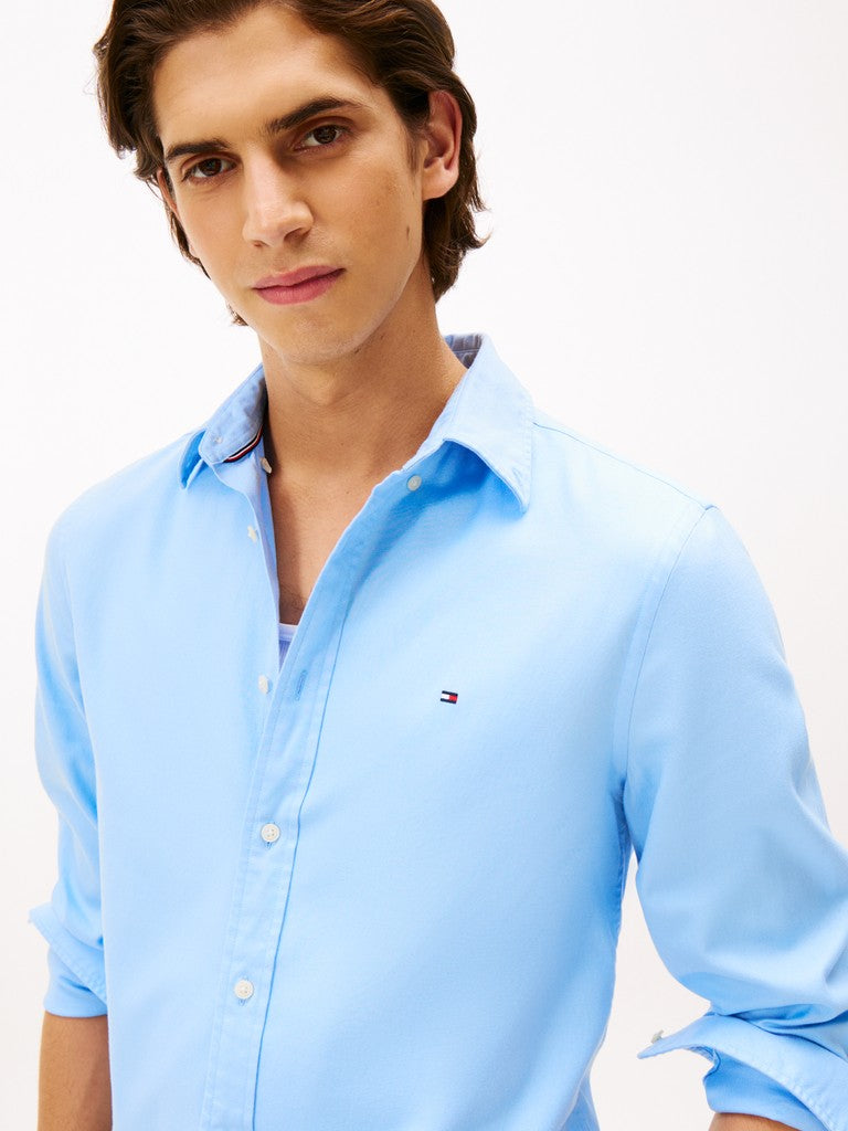 A man wearing a light blue Tommy Hilfiger button-down shirt, shown from the chest up against a plain white background.