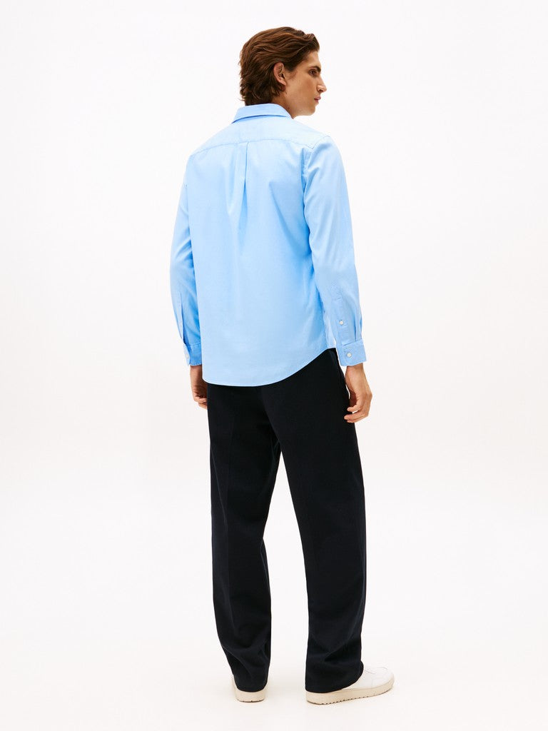 A man shown from the back wearing a light blue long-sleeve button-up shirt, dark navy trousers, and white sneakers, photographed against a clean white studio background.