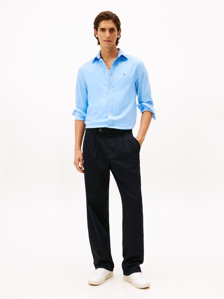 A man modeling a light blue long-sleeve button-down shirt with rolled-up sleeves, paired with dark navy trousers and white sneakers, against a plain white studio background.