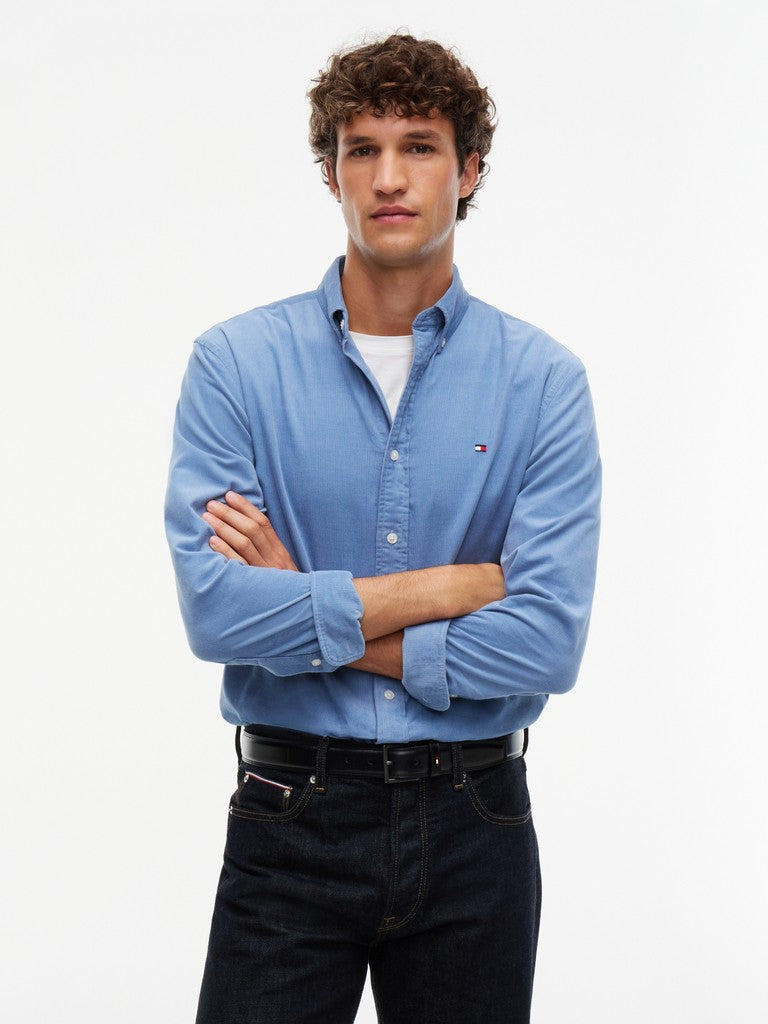 A man wearing a light blue long-sleeve button-down shirt by Tommy Hilfiger, paired with dark jeans and a white undershirt, standing against a plain white background.