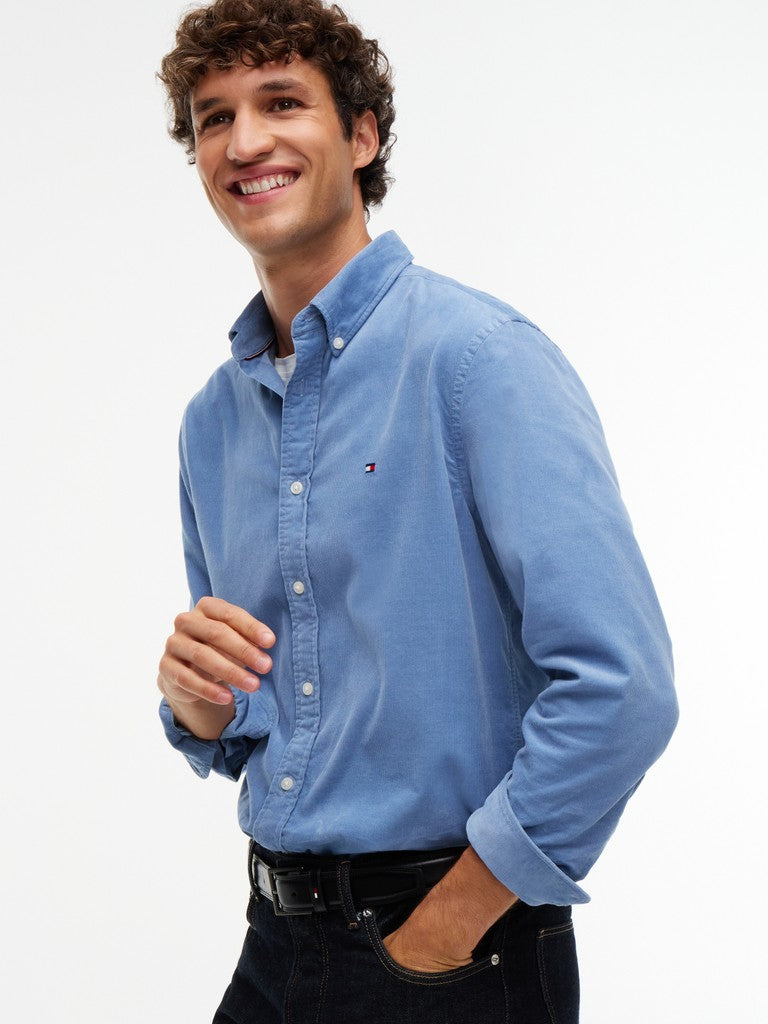 A smiling man wearing a light blue long-sleeve button-down shirt with a small embroidered Tommy Hilfiger flag logo on the chest, paired with dark jeans against a plain white background.