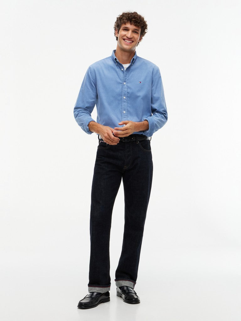 A man modeling a light blue long-sleeve button-down shirt and dark navy jeans, standing against a plain white studio background.
