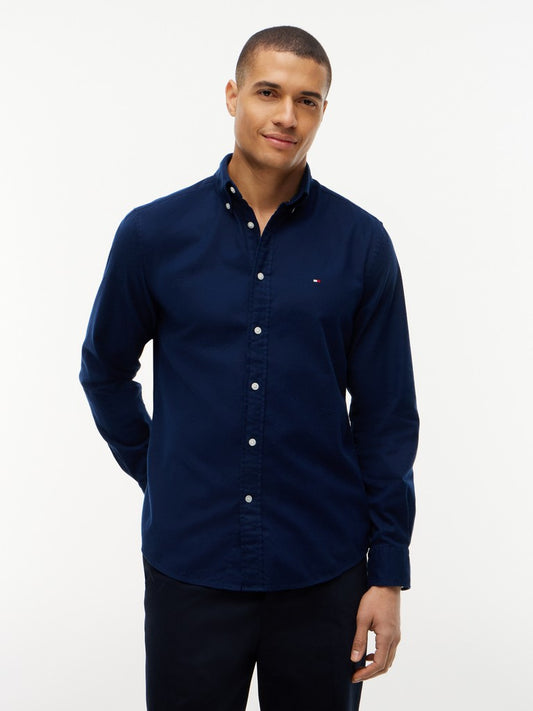 A man wearing a navy blue long-sleeve button-down shirt with a small embroidered Tommy Hilfiger flag logo on the chest, paired with dark trousers, against a plain white studio background.