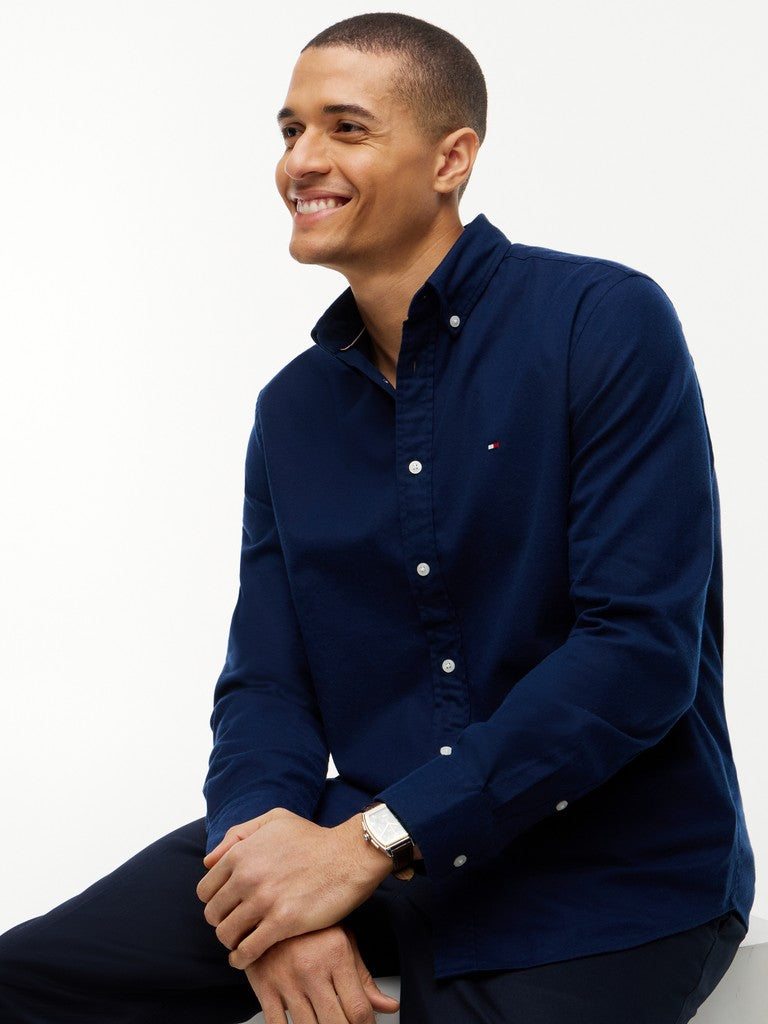 A man wearing a navy blue long-sleeve button-down shirt and matching navy trousers, seated against a plain white studio background.