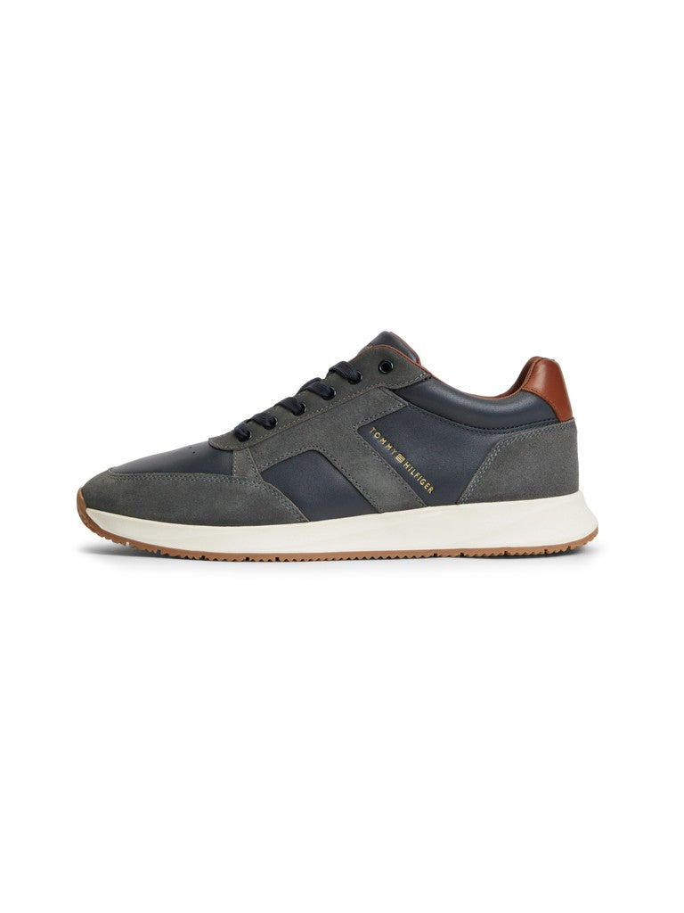 A low-top sneaker by Tommy Hilfiger, featuring a dark gray suede and navy blue leather upper, a thick white midsole, and a brown heel tab, shown from a side profile on a white background.
