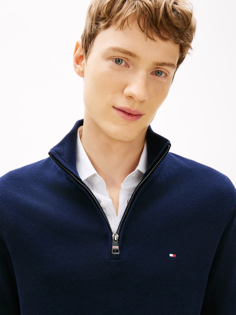 A close-up of a man wearing a navy blue quarter-zip sweater by Tommy Hilfiger over a white collared shirt, against a clean white background.