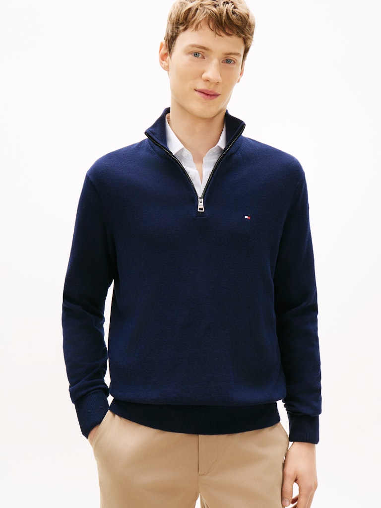 Man wearing a navy blue sweater with a Tommy Hilfiger flag logo on the chest and a white shirt underneath on a light gray background