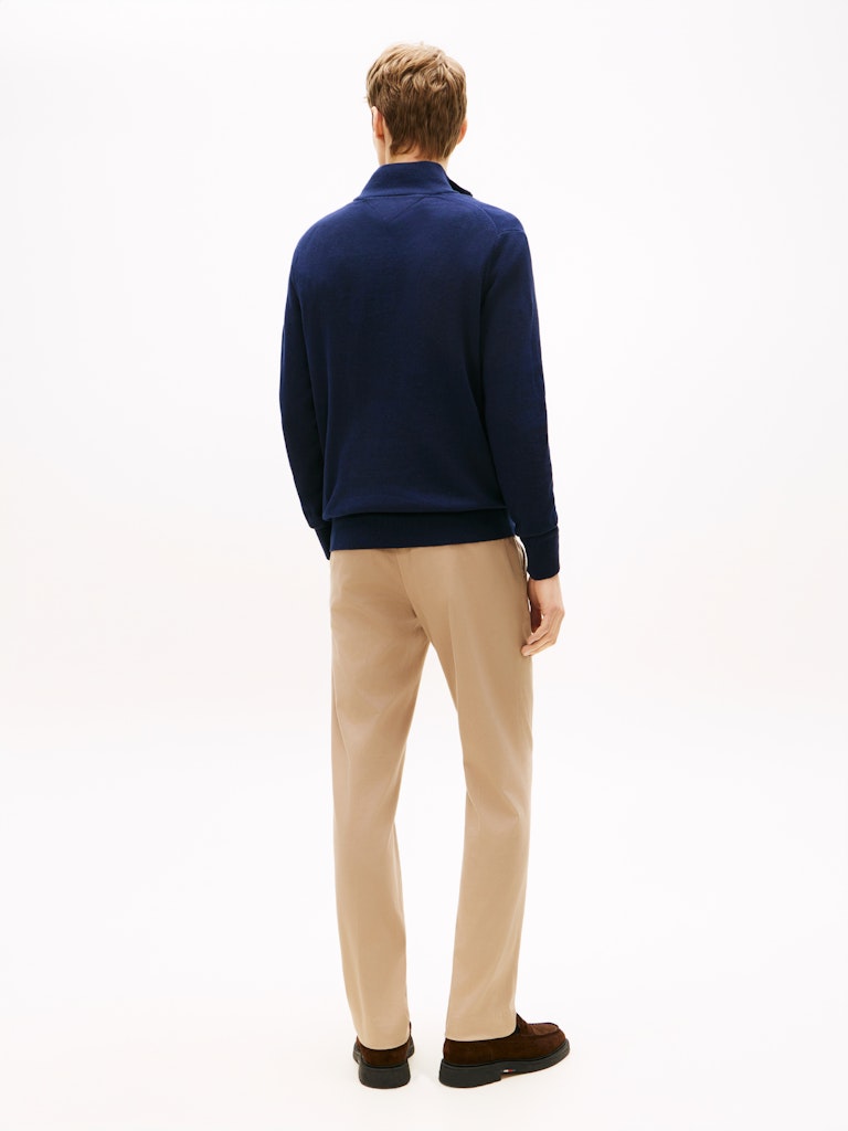 A man viewed from behind wearing a navy blue knit quarter-zip sweater, beige trousers, and dark brown loafers, standing against a plain white studio background.