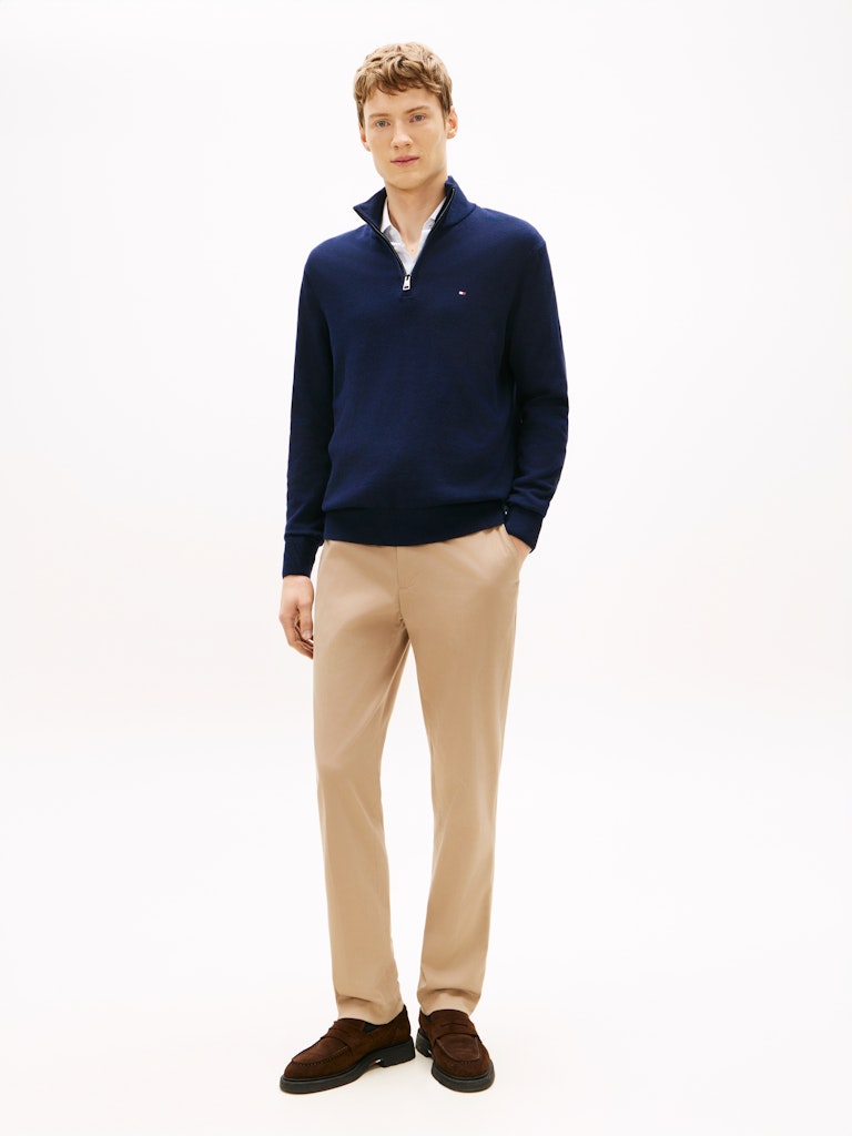 Man wearing a navy blue quarter-zip sweater by Tommy Hilfiger over a white collared shirt and cream trousers, against a clean white background.