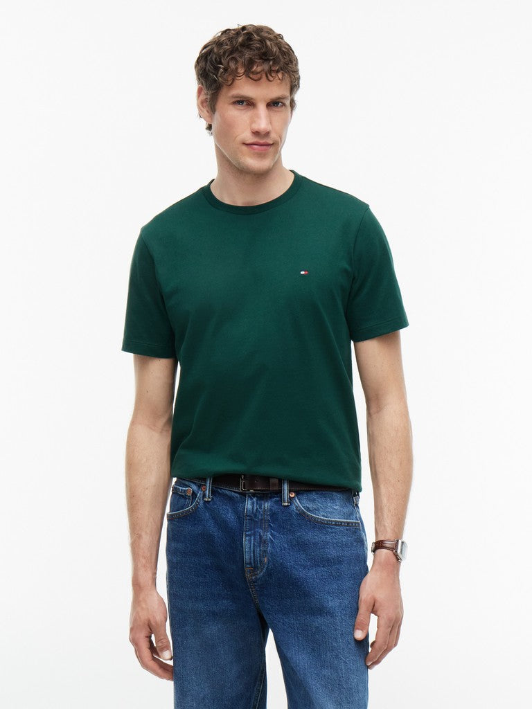 A man wearing a dark green short-sleeve crewneck t-shirt with a small embroidered Tommy Hilfiger flag logo on the left chest, paired with blue jeans, against a plain white studio background.