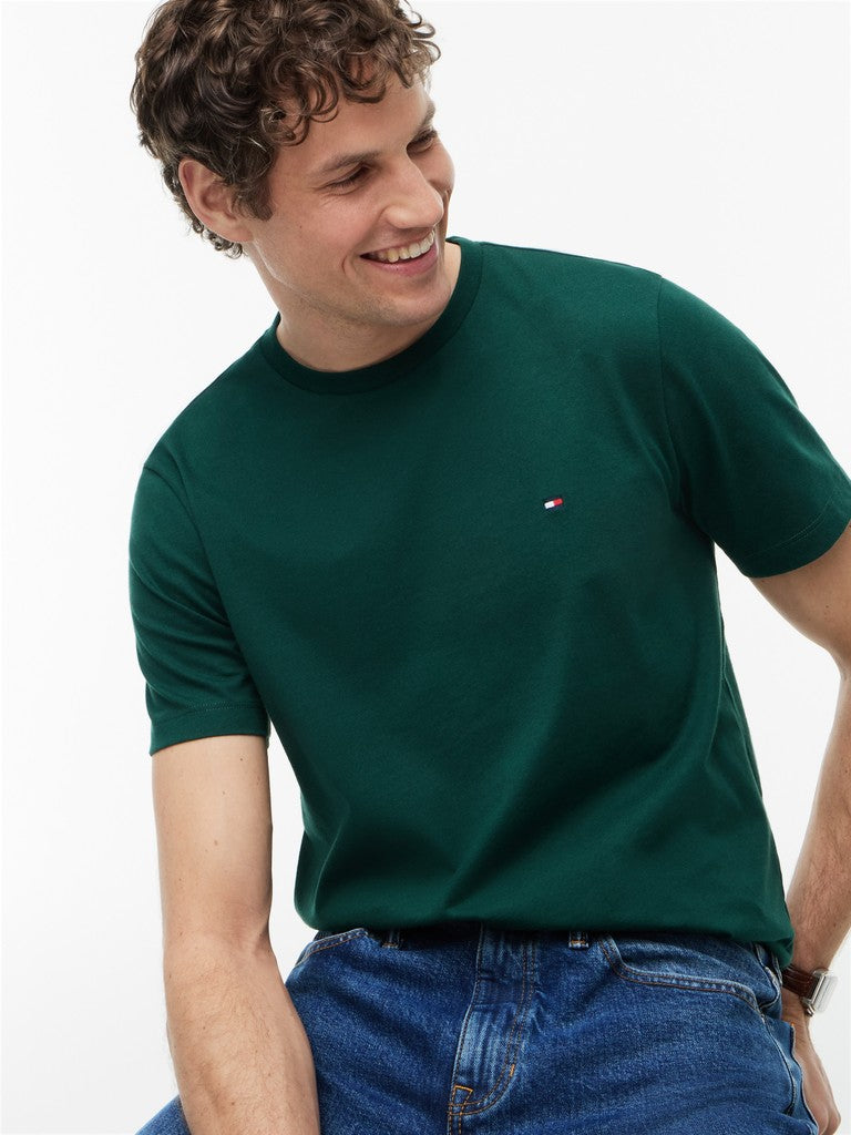 A man wearing a dark green short-sleeve crewneck t-shirt with a small embroidered Tommy Hilfiger flag logo on the left chest, paired with blue jeans, against a plain white background.