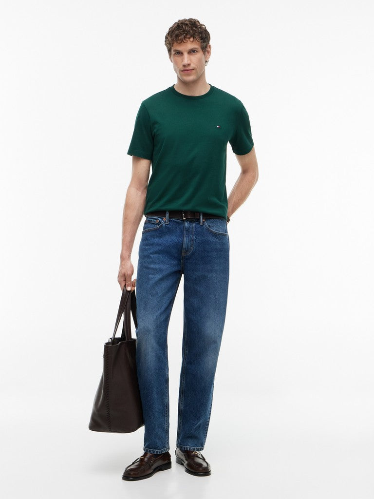 A man modeling a dark green short-sleeve crewneck t-shirt, blue jeans, and dark brown leather loafers, holding a black tote bag against a plain white studio background.