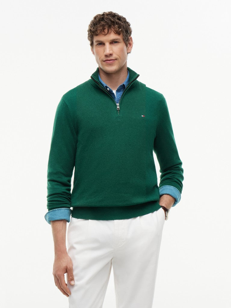 A man wearing a dark green quarter-zip sweater with a small embroidered Tommy Hilfiger flag logo on the chest, layered over a blue collared shirt and paired with white trousers, against a plain white background.