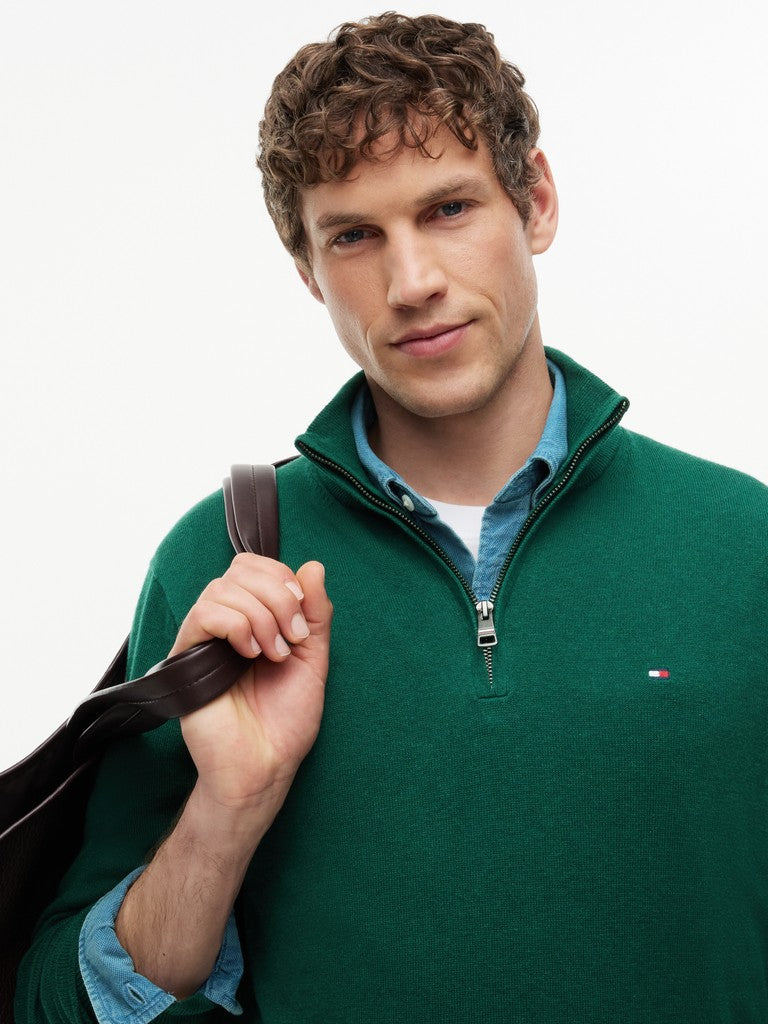 A man wearing a dark green quarter-zip sweater by Tommy Hilfiger, layered over a light blue button-down shirt and a white undershirt, holding a dark brown bag strap against a plain white background.
