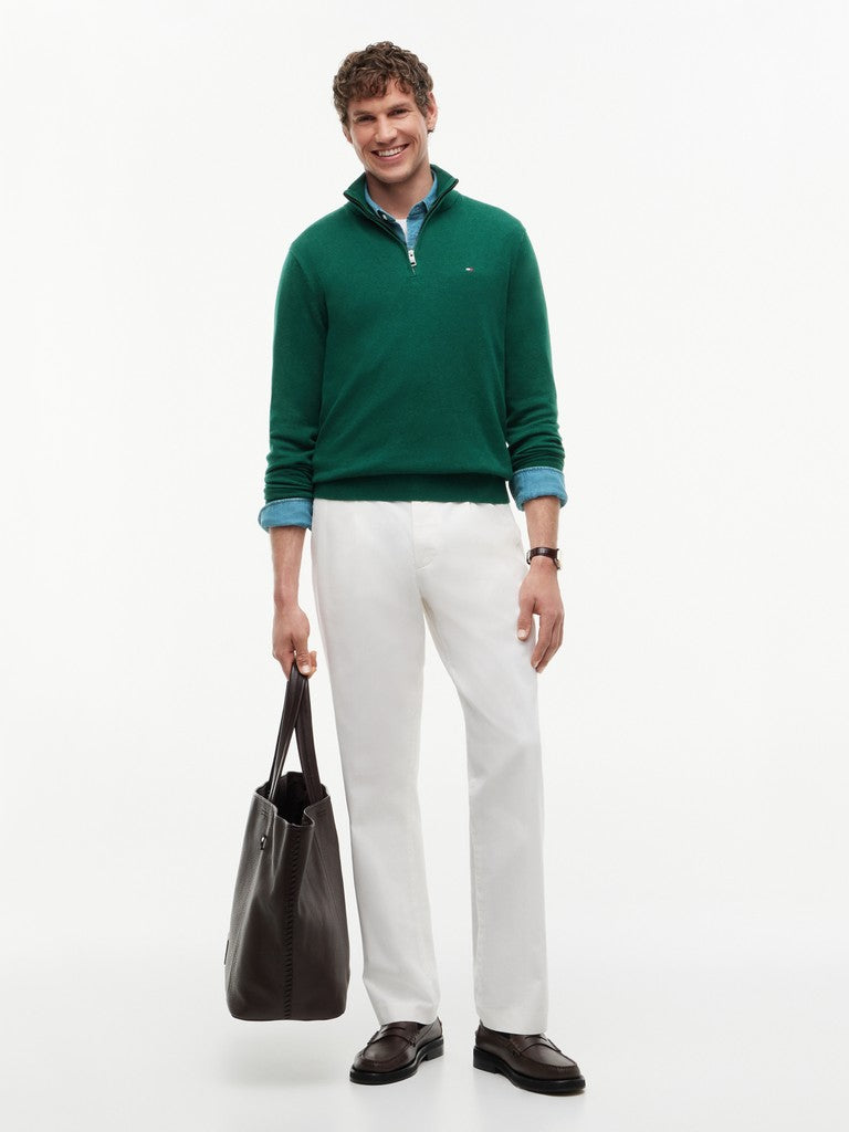A man modeling a green quarter-zip sweater with a blue collar lining, paired with white trousers and holding a black tote bag against a plain white studio background.