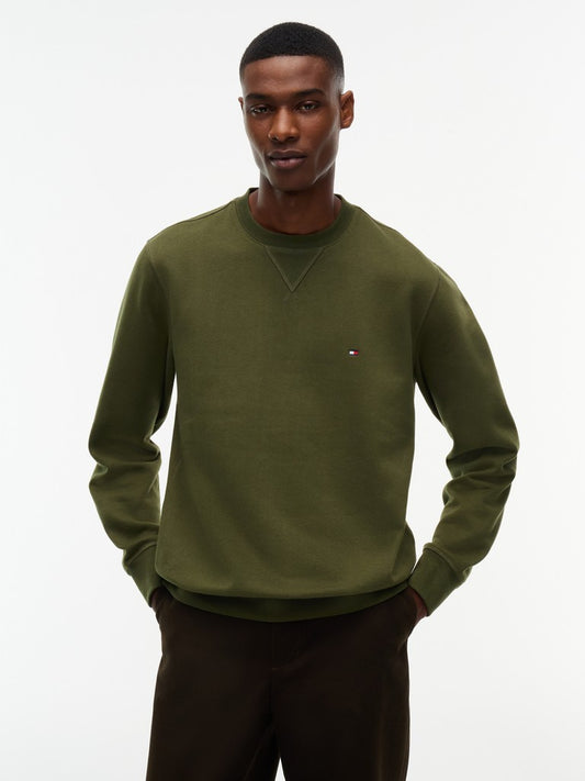 A man wearing an olive green crewneck sweatshirt with a small embroidered Tommy Hilfiger flag logo on the left chest, paired with dark brown pants, against a plain white studio background.