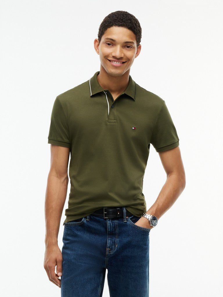 A man wearing an olive green short-sleeve polo shirt with a small embroidered Tommy Hilfiger flag logo on the chest, paired with blue jeans, against a plain white studio background.