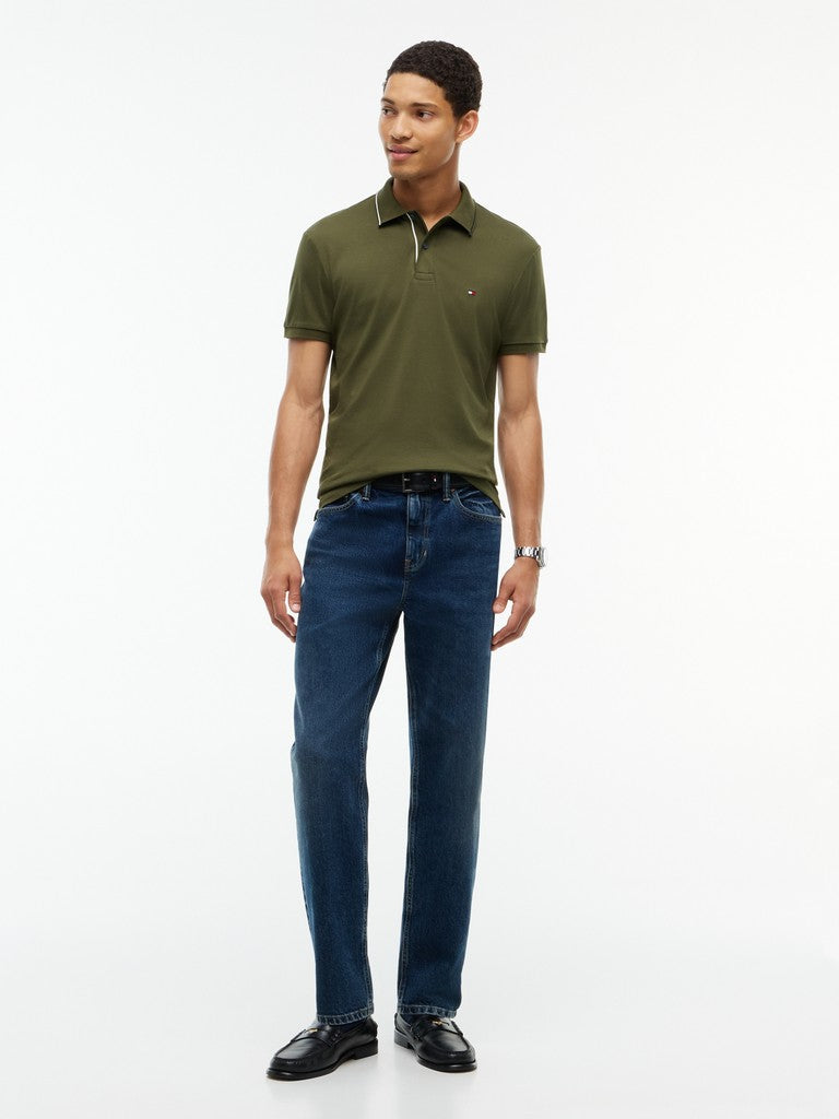 A man modeling a short-sleeve olive green polo shirt with a small embroidered Hilfiger logo on the chest, paired with dark blue jeans and black shoes, against a plain white studio background.
