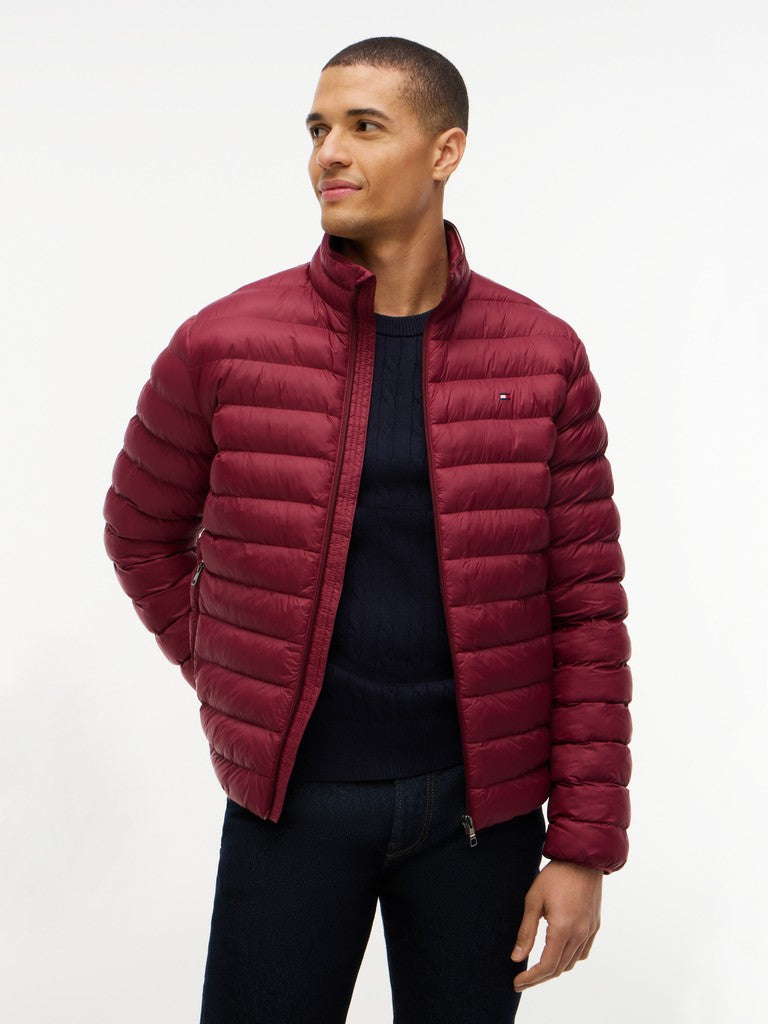 A man wearing a burgundy quilted puffer jacket over a dark navy sweater and dark jeans, standing against a plain white studio background.