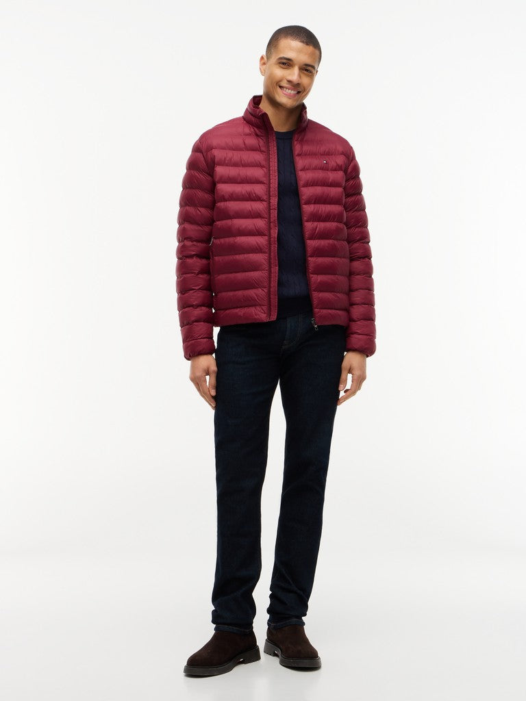 A man modeling a dark red puffer jacket over a navy blue sweater and dark jeans, standing against a plain white studio background.