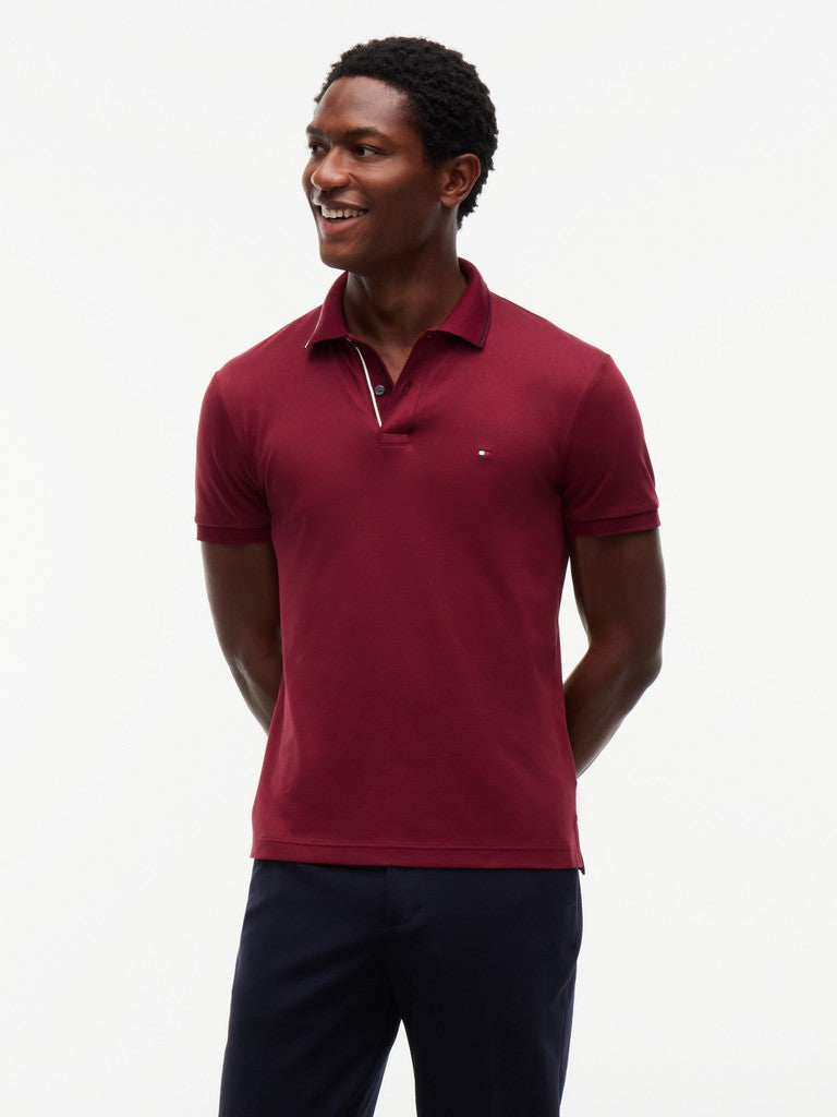 A man wearing a short-sleeve polo shirt in burgundy with a small white embroidered logo on the chest, paired with dark trousers, photographed against a plain white studio background.