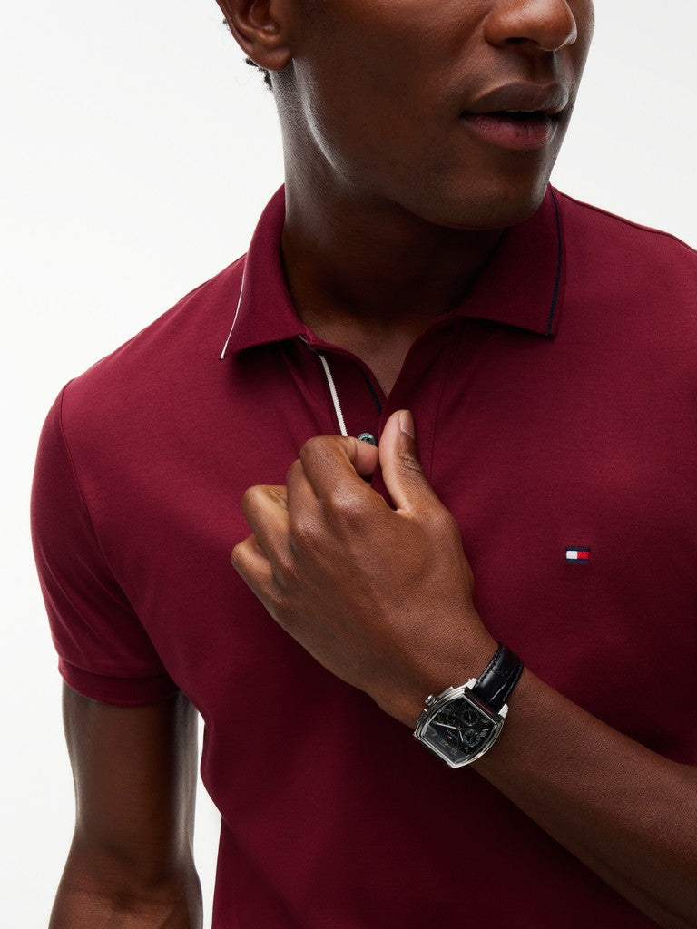 A close-up of a man wearing a short-sleeve polo shirt in deep burgundy. The shirt features a small embroidered Tommy Hilfiger flag logo on the left chest and a white tipping detail on the collar and placket.
