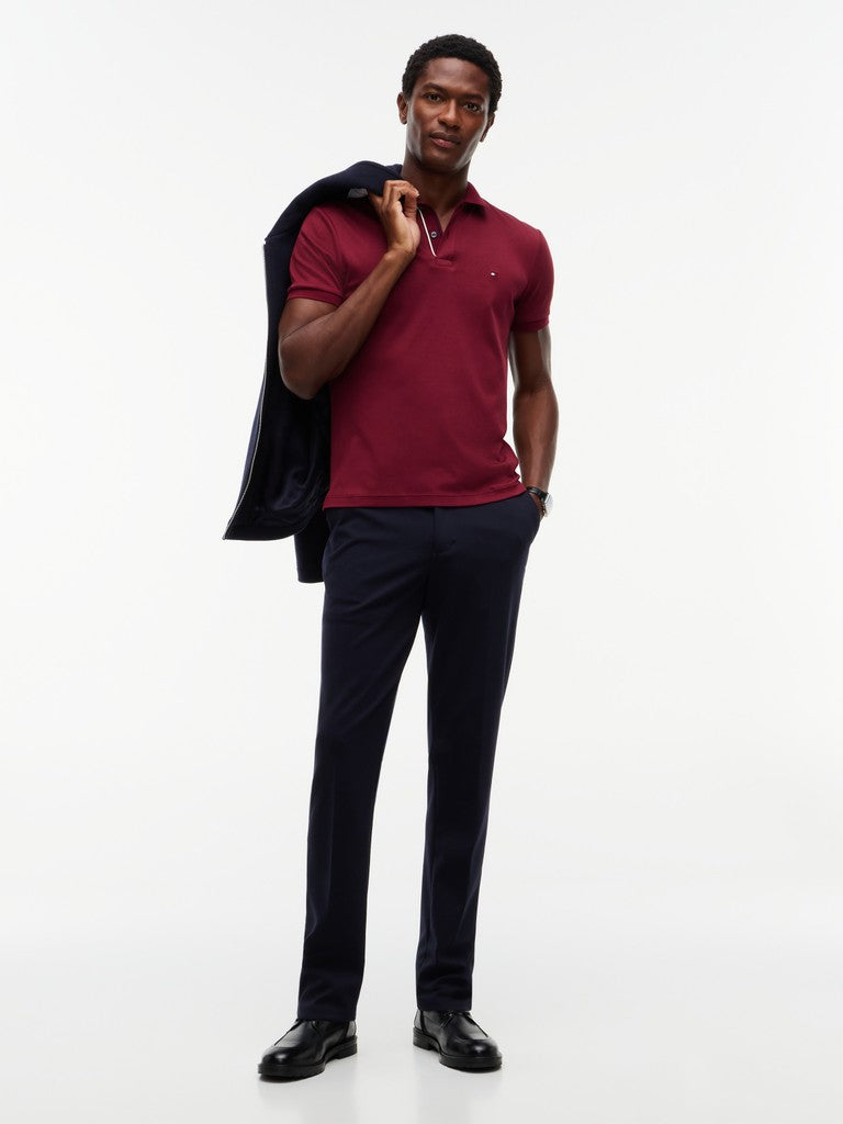 A man modeling a maroon short-sleeve polo shirt, navy trousers, and black dress shoes, with a navy jacket draped over his shoulder, against a plain white studio background.