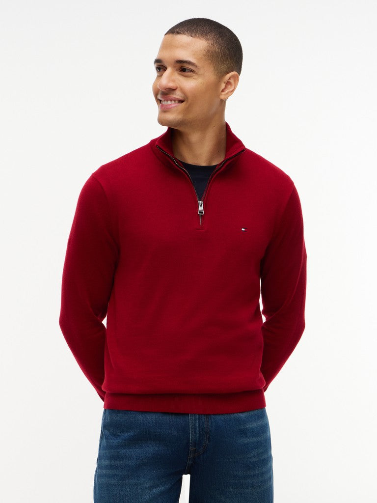 A close-up of a man wearing a red quarter-zip sweater by Tommy Hilfiger over a navy T-shirt and blue jeans, against a clean white background.