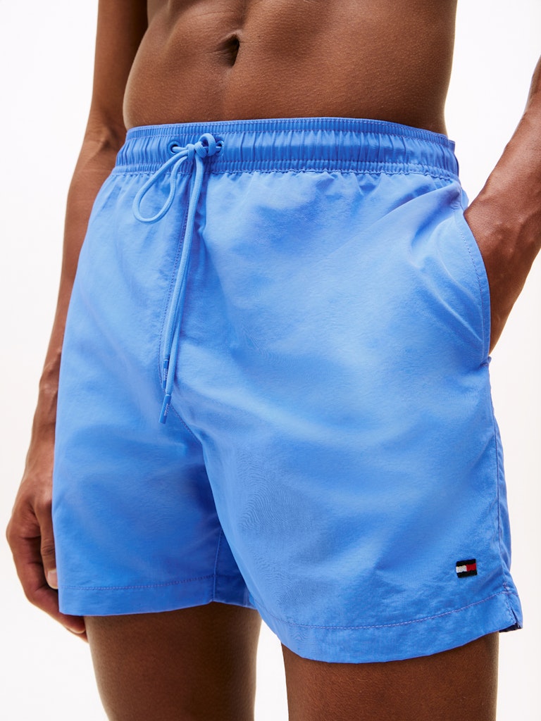 A close-up of a model wearing light blue swim trunks with a drawstring waist and a small Tommy Hilfiger flag logo near the hem.