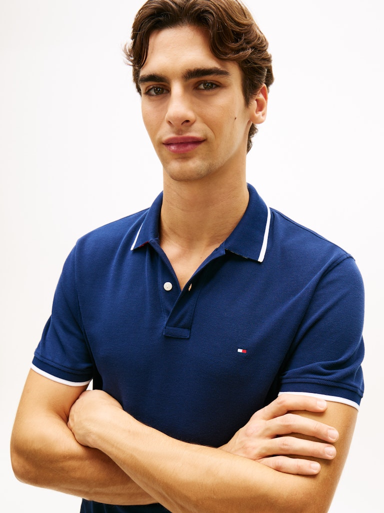 Man wearing a blue Tommy Hilfiger polo shirt with a white collar against a light gray background