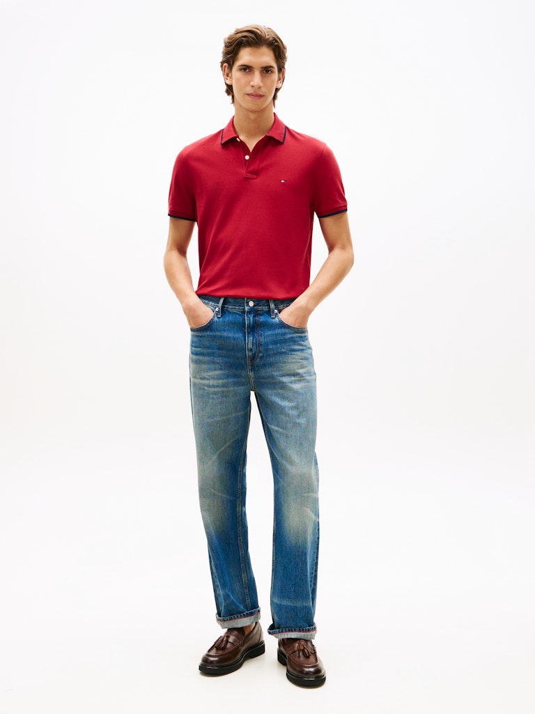 Man wearing a red Tommy Hilfiger polo with blue jeans and brown shoes against a white background