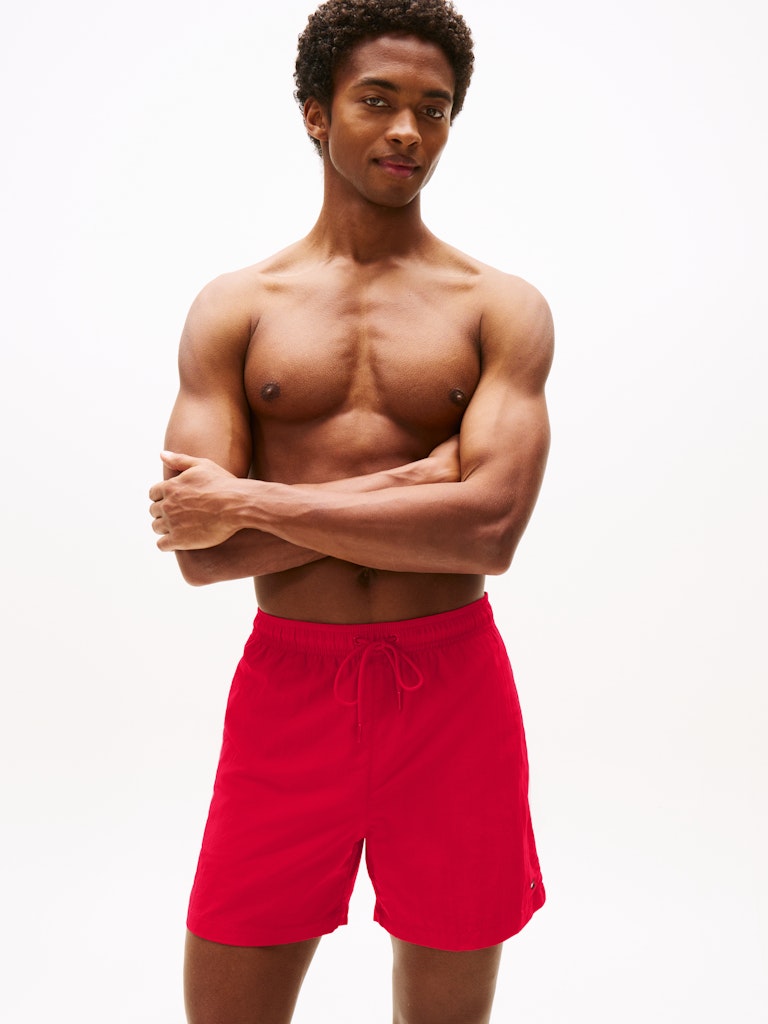 A shirtless man modeling bright red swim trunks against a clean white studio background.