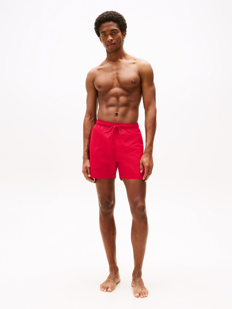 A shirtless man wearing bright red swim trunks stands against a plain white studio background.