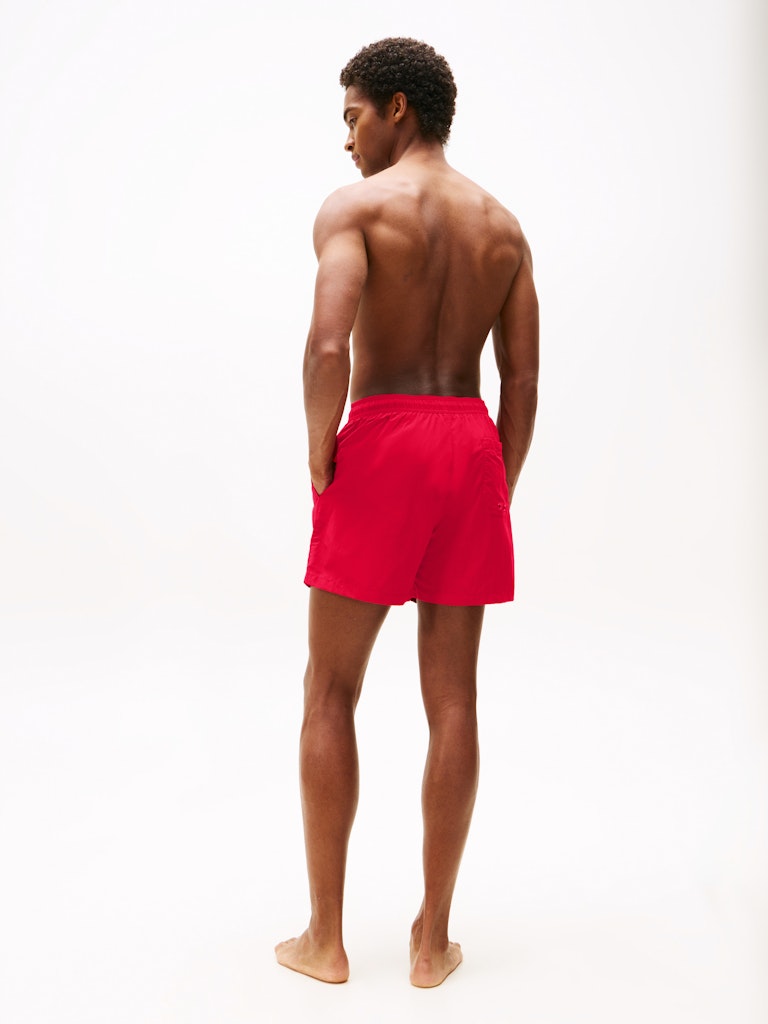 A man shown from behind wearing bright red swim shorts, standing barefoot against a plain white studio background.